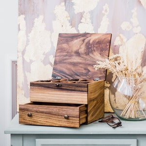 Pu&ograve; includere: Una scatola di legno con un coperchio incernierato e un cassetto. La scatola &egrave; realizzata in legno scuro con una venatura naturale. Il coperchio &egrave; aperto e l'interno della scatola &egrave; visibile. La scatola &egrave; appoggiata su un tavolo blu chiaro.