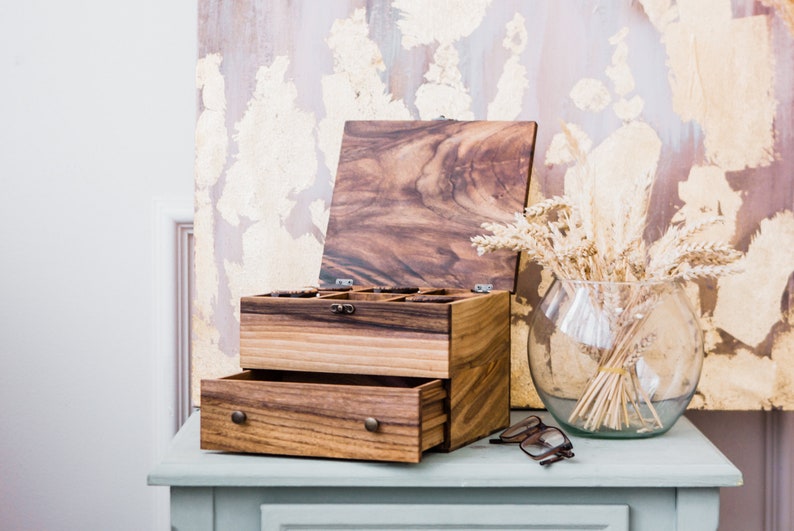 Pu&ograve; includere: Una scatola di legno con un coperchio incernierato e un cassetto. La scatola &egrave; realizzata in legno scuro con una venatura naturale. Il coperchio &egrave; aperto e l'interno della scatola &egrave; visibile. La scatola &egrave; appoggiata su un tavolo blu chiaro.