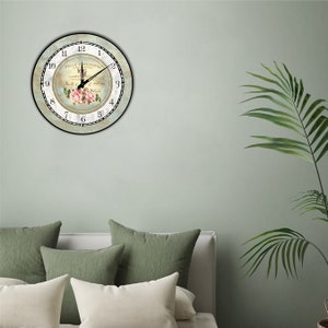 Pu&ograve; includere: Un orologio da parete rotondo con un design floreale. Il quadrante dell'orologio presenta un'estetica vintage con il testo "BELLE JARDINIERE" e numeri romani. Le lancette dell'orologio segnano l'1:00. L'orologio &egrave; montato su una parete verde chiaro.