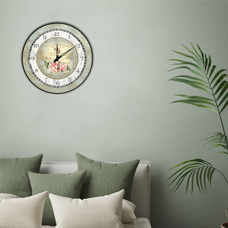 Pu&ograve; includere: Un orologio da parete rotondo con un design floreale. Il quadrante dell'orologio presenta un'estetica vintage con il testo "BELLE JARDINIERE" e numeri romani. Le lancette dell'orologio segnano l'1:00. L'orologio &egrave; montato su una parete verde chiaro.