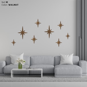 Puede incluir: Juego M, decoración de pared de estrellas color nogal. Nueve estrellas de madera de diferentes tamaños están montadas en una pared blanca sobre un sofá gris. Las estrellas tienen un acabado marrón cálido, creando una estética moderna.