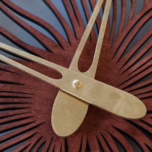 May include: Close-up of a gold-colored clock hand with a circular, metallic accent. The hand is set against a background of dark red wood with a radial pattern.