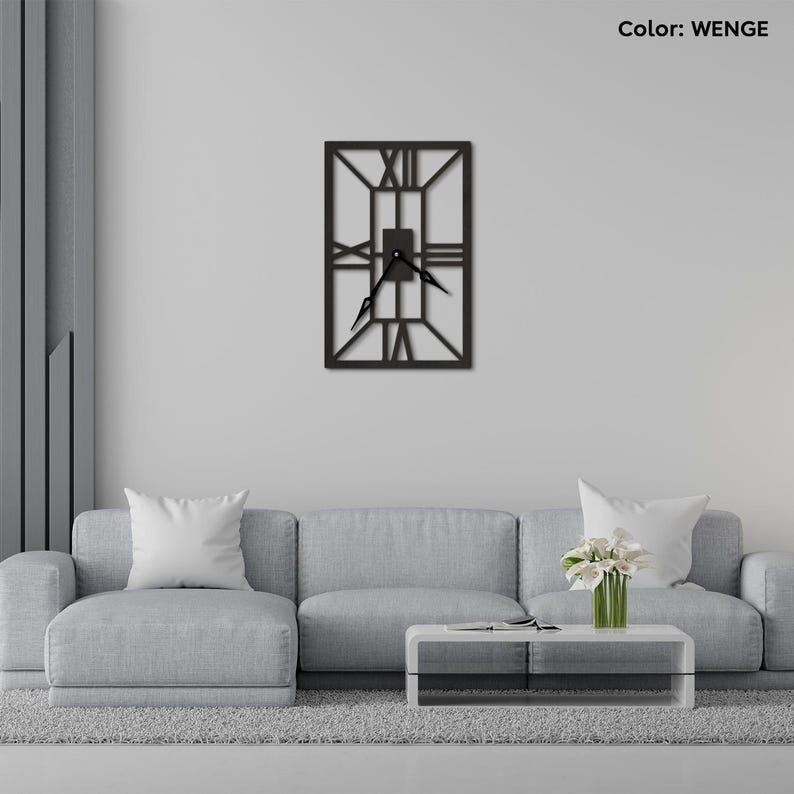 May include: A black metal wall clock with Roman numeral markings and a square frame. The clock is hanging on a white wall above a gray couch and a white coffee table.