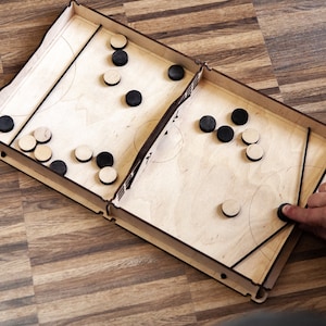 May include: A wooden tabletop game with a light brown finish. The game features a rectangular playing surface with circular cutouts and black and light brown game pieces. A hand is seen playing the game.