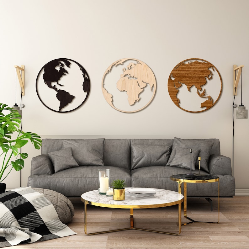 May include: Three wooden world map wall decor pieces in a living room setting. The maps are in different wood tones, with one being black, one being natural wood, and one being a darker wood tone. The maps are hanging on a white wall above a gray couch and a white marble coffee table with a gold metal frame.