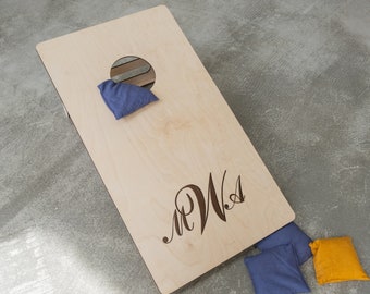 May include: A wooden cornhole game with a personalized monogram "M.W.S." and two blue bean bags. A yellow bean bag is also visible on the ground.