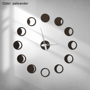 Könnte beinhalten: Eine moderne Wanduhr mit dunkelbraunem Zifferblatt und weißen Zeigern. Das Zifferblatt besteht aus zwölf kreisförmigen Mondphasenformen. Der Text "Color: palisander" befindet sich in der oberen linken Ecke.