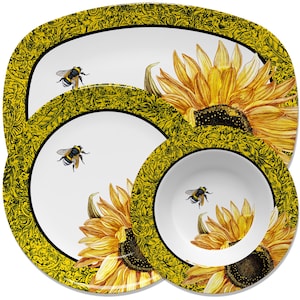 May include: A set of three white ceramic plates with a sunflower and bee design. The plates have a yellow rim with a black floral pattern. The largest plate is oval shaped, the middle plate is round, and the smallest plate is a bowl.
