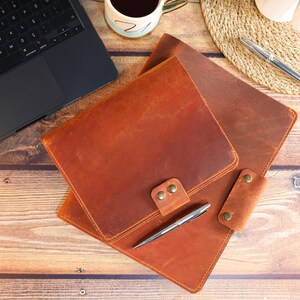 May include: Two brown leather notebook covers with a silver pen on top of the larger cover. The covers have a flap closure and a metal button.