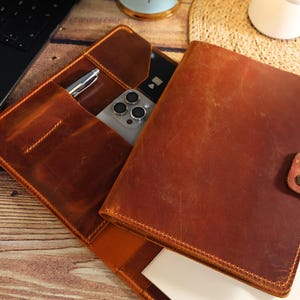 May include: A brown leather notebook with a built-in phone pocket and pen holder. The notebook has a flap closure and a stitched edge.