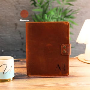 May include: A brown leather notebook cover featuring a vertical strap with two brass-colored metal snaps. The front cover is engraved with the letter A and the name Adam. The leather has a smooth, matte finish with visible stitching.
