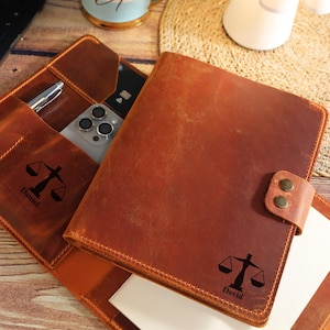 May include: A brown leather portfolio organizer featuring a snap closure, interior pockets for a smartphone and pen, and a laser-engraved scale of justice symbol with the names Daniel and David printed on the exterior and interior surfaces.