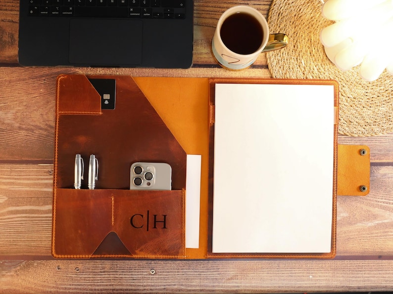 May include: A brown leather portfolio with a flap closure, two pen slots, a phone pocket, and a card slot. The portfolio has a personalized monogram "C|H" and a white sheet of paper inside.