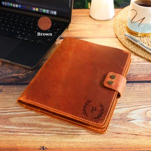 May include: A brown leather notebook cover featuring a snap closure with two brass-colored buttons and a laser-engraved laurel wreath design surrounding the letter P on the front cover, showcasing a smooth, matte texture.