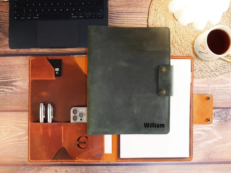 May include: A brown leather portfolio with a green leather flap. The flap has a personalized inscription that reads "William". The portfolio has a pocket for pens and a pocket for a phone. The portfolio is open to reveal a white sheet of paper.
