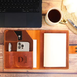 May include: A tan leather notebook cover featuring a personalized monogram with the letter D and the name Daniel. The interior includes slots for a smartphone, a credit card, a pen, and a notebook, with a snap closure on the right side.