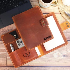 May include: A brown leather notebook cover featuring a snap closure, the name Kevin embossed on the front, and an interior pocket with a K and antler graphic. The organizer includes slots for a smartphone, a pen, and a card, with a white notepad.