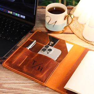 May include: A brown leather notebook cover featuring a pocket with the monogram T/H, containing a silver pen, a smartphone, and a black card. The cover is open, revealing a blank white notebook page on the right side, resting on a wooden desk surface.
