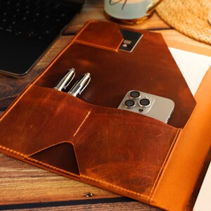 May include: A brown leather portfolio with a flap closure, featuring a pocket for a phone and two pen slots. The portfolio is open to reveal a notepad and a white paper.