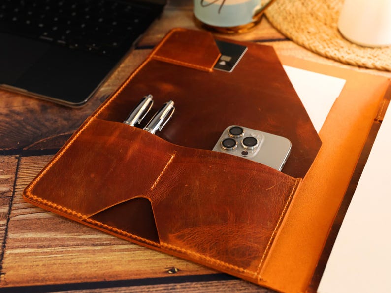 May include: A brown leather portfolio with a flap closure, featuring a pocket for a phone and two pen slots. The portfolio is open to reveal a notepad and a white paper.