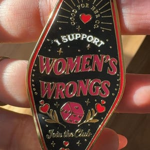K&ouml;nnte beinhalten: Goldener Schl&uuml;sselanh&auml;nger mit einem schwarzen Emaille-Anh&auml;nger in Form eines Motel-Schilds. Der Anh&auml;nger hat roten und wei&szlig;en Text, der "Good for Her Co. I Support Women's Wrongs. Join the Club." lautet. Der Anh&auml;nger zeigt au&szlig;erdem ein rotes Herz, einen roten W&uuml;rfel mit Herzen und goldene Sterne.