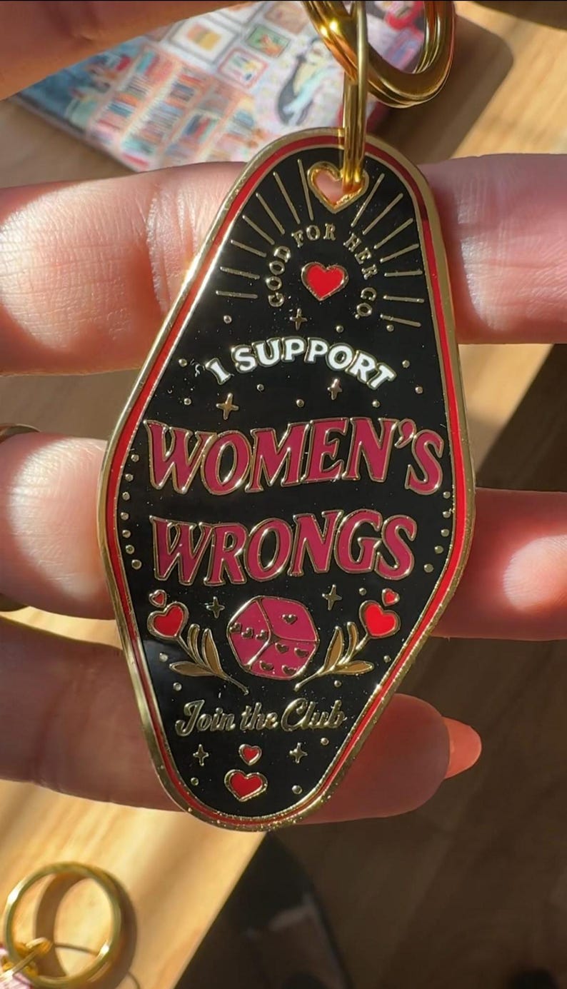 K&ouml;nnte beinhalten: Goldener Schl&uuml;sselanh&auml;nger mit einem schwarzen Emaille-Anh&auml;nger in Form eines Motel-Schilds. Der Anh&auml;nger hat roten und wei&szlig;en Text, der "Good for Her Co. I Support Women's Wrongs. Join the Club." lautet. Der Anh&auml;nger zeigt au&szlig;erdem ein rotes Herz, einen roten W&uuml;rfel mit Herzen und goldene Sterne.