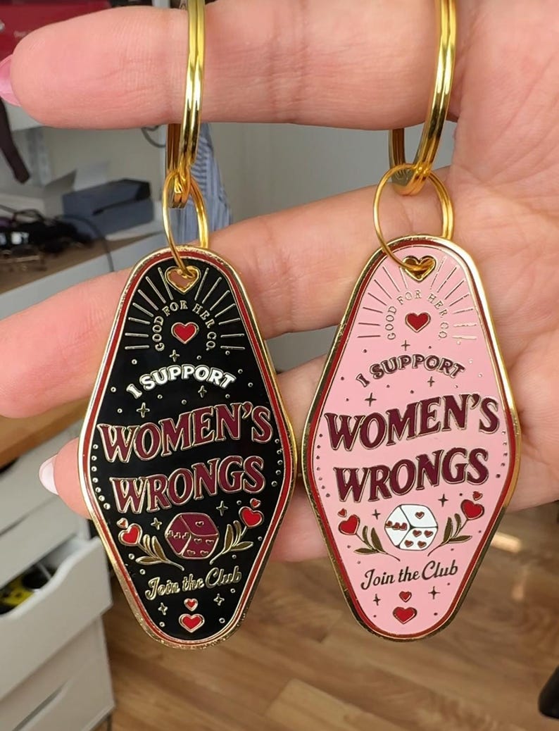 K&ouml;nnte beinhalten: Zwei Schl&uuml;sselanh&auml;nger mit Motel-Schl&uuml;ssel-Design. Beide Schl&uuml;sselanh&auml;nger sind golden mit schwarzer und rosa Emaille. Der Text auf den Schl&uuml;sselanh&auml;ngern lautet "Good for Her Co." und "I Support Women's Wrongs." Die Schl&uuml;sselanh&auml;nger haben auch ein rotes Herz, einen W&uuml;rfel und den Text "Join the Club."