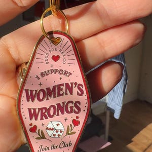 K&ouml;nnte beinhalten: Rosafarbener Schl&uuml;sselanh&auml;nger aus Metall mit einem Motel-Schild-Design. Es steht "Good For Her Co." und "I Support Women's Wrongs."  Es hat auch ein rotes Herz und W&uuml;rfel mit Herzen darauf. Der Text unten lautet "Join the Club."