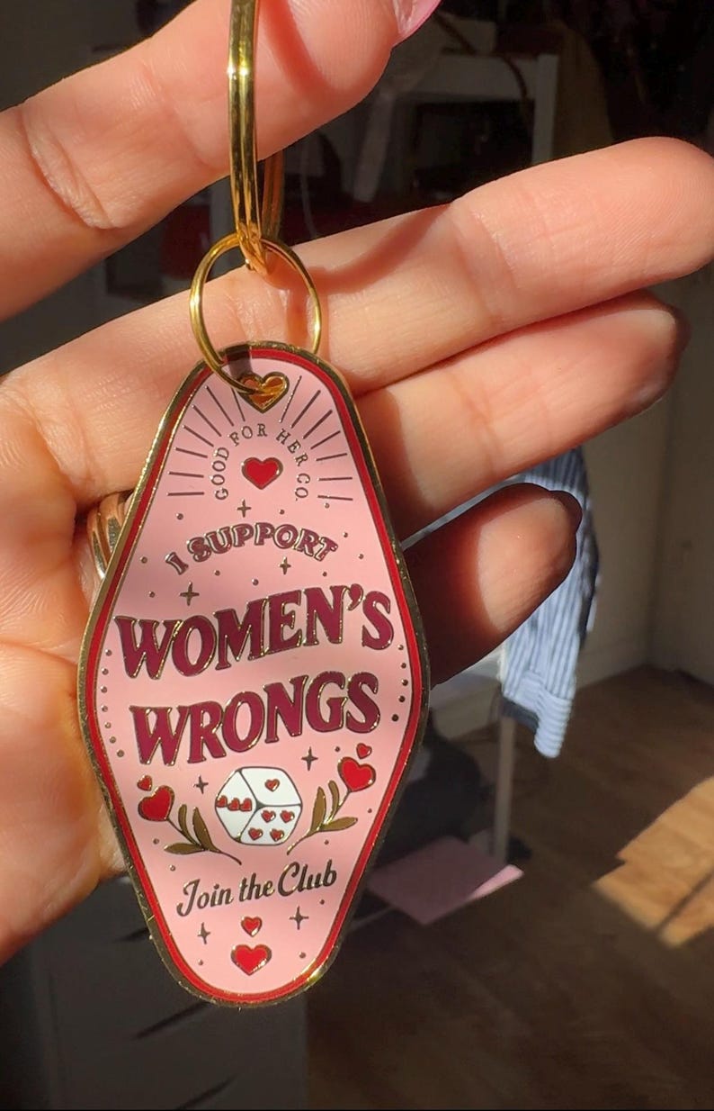 K&ouml;nnte beinhalten: Rosafarbener Schl&uuml;sselanh&auml;nger aus Metall mit einem Motel-Schild-Design. Es steht "Good For Her Co." und "I Support Women's Wrongs."  Es hat auch ein rotes Herz und W&uuml;rfel mit Herzen darauf. Der Text unten lautet "Join the Club."