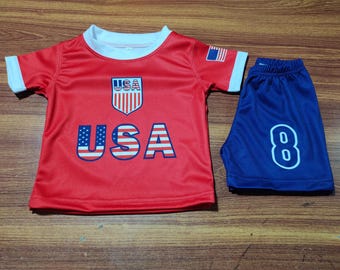 May include: A red and blue USA soccer uniform set. The short-sleeved shirt has a white collar and cuffs, with "USA" in large letters and a shield logo. The blue shorts feature the number "8".