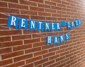 May include: A blue banner with white text spelling "RENTNER 2025" and "HANS" hangs against a red brick wall. The banner has a heart symbol between the names and the year. The letters are in a serif font.