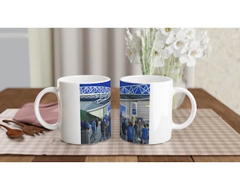 May include: Two white ceramic mugs with a blue and white illustration of a stadium and fans. The mugs are on a wooden table with a light brown placemat.