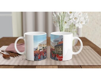 May include: Two white ceramic mugs with a colorful illustration of a stadium and fans in red shirts. The mugs are on a checkered tablecloth.