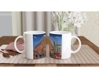 May include: Two white ceramic mugs with a wraparound illustration of a stadium at night. The artwork features a crowd of people, stadium architecture, and a dark blue sky. The mugs are on a wooden table.