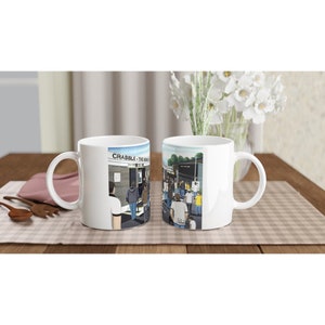 May include: Two white ceramic mugs with a colorful illustration of a crowd of people entering a stadium. The text "CRABBLE - THE HOME OF THE..." is visible on the mug.