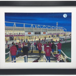 May include: A framed art print depicting a night scene at a football stadium with a full moon in the dark blue sky. Fans wearing red jerseys with numbers walk down stairs toward the stadium entrance. The text "Lichfield Stadium" is visible.