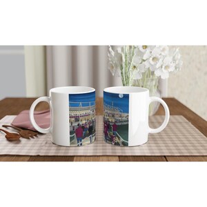 Peut inclure: Deux mugs en céramique blanche avec une illustration d'un stade la nuit. Les mugs sont posés sur une table en bois avec un set de table à carreaux. Un vase de fleurs blanches et des ustensiles en bois sont en arrière-plan.