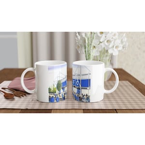 Puede incluir: Dos tazas de cerámica blanca con una ilustración azul y blanca de un estadio en el lateral. Las tazas están sobre una mesa de madera con un mantel a cuadros.