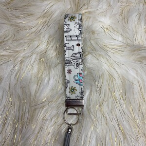 May include: A white key chain with a black and white pattern of teacups, flowers, and cars. The key chain has a silver metal ring and a gray tassel.