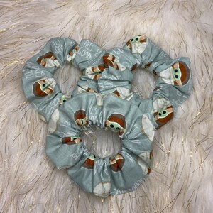 May include: Three light blue scrunchies with a repeating pattern of a green alien character with the text "This is the way" on a white background.