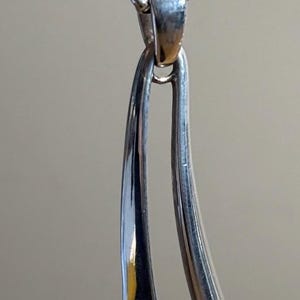 May include: A silver pendant necklace with a unique, elongated design. The pendant features two curved, polished silver elements, connected at the top by a chain. The pendant has two inset stones, one light pink and one light blue.