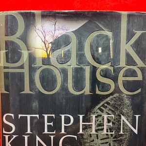 May include: A black hardback book titled "Black House" by Stephen King and Peter Straub. The book cover features a dark silhouette of a house with a tree in front of it. The authors are listed as "Authors of The Talisman" and the book is described as "A Novel".