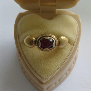 Pode incluir: Um anel de ouro com uma pedra preciosa vermelha, apresentado numa caixa de joias em forma de coração creme. O anel apresenta uma pedra central oval e dois detalhes redondos. O interior da caixa é de veludo amarelo.