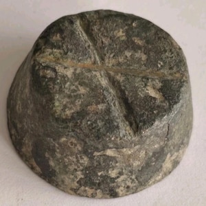 May include: A circular, conical metal weight with a weathered, dark grey and mottled surface. The top flat surface features a deeply incised cross mark that extends across the diameter, showing signs of oxidation and age-related surface wear.