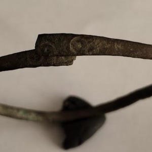 Peut inclure: Un bracelet en métal terni d'aspect sombre et vieilli. Le bracelet présente un design simple et incurvé avec un fermoir et un petit élément sombre en forme de cœur. Le métal montre des signes d'usure et de vieillissement.