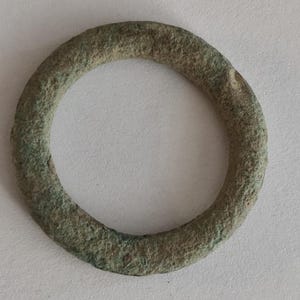 May include: A circular ring with a weathered, textured surface in shades of muted green, brown, and gray. The object features a consistent, rounded cross-section and a rough, uneven patina throughout its entire circumference, showing signs of age.