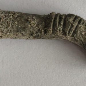 May include: A weathered, gray, metallic hook-shaped object with a textured surface. The item features a series of parallel ridges or grooves etched into the metal at both the straight end and the curved end, showing signs of age and oxidation.