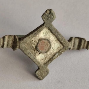 Op de afbeelding: Een metalen ring met een ruitvormig voorvlak met een getextureerd, verweerd grijs oppervlak. Het midden bevat een cirkelvormige, roodbruine inleg omringd door een witte rand. De ring wordt geflankeerd door twee decoratieve, geribbelde uitsteeksels.