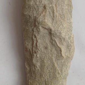 Pode incluir: Uma pedra de cor bege claro, de forma aproximadamente retangular, com uma superfície texturizada. A pedra tem um aspeto natural e envelhecido, com variações subtis de cor e pequenas reentrâncias.
