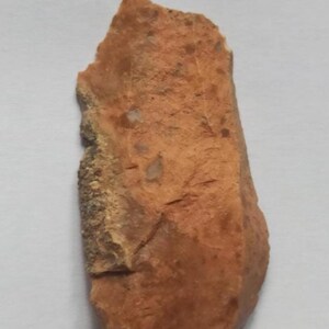 May include: A small, irregularly shaped piece of reddish-brown stone with a rough, textured surface. The stone has a lighter-colored area and a darker, more textured edge. The overall appearance suggests a natural, possibly ancient artifact.